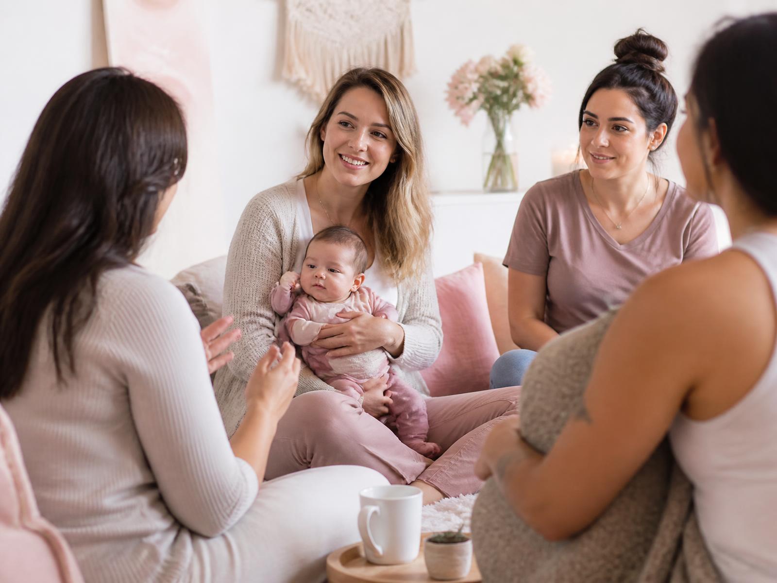 Plusieurs mamans assises ensemble et s’écoutant dans un salon lumineux, l’une d’elles portant son bébé